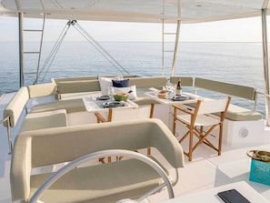 A boat deck with a table set for two, two chairs, a small table with a bottle, and a bowl of food.