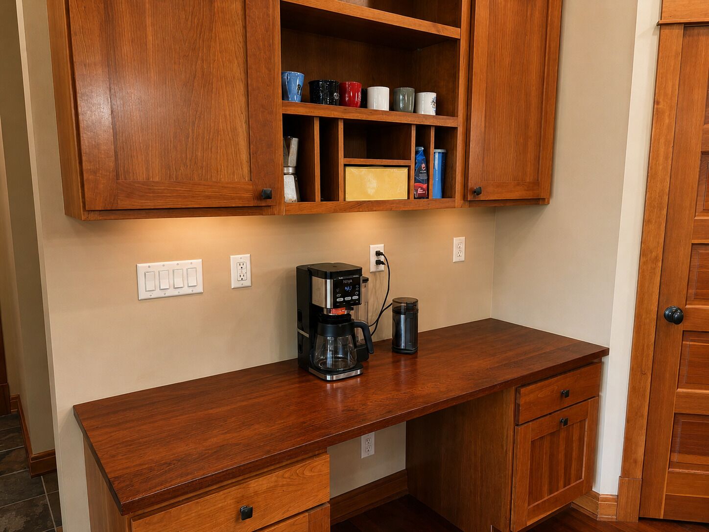 A wooden kitchen counter with a coffee maker and a kettle, a cabinet above with various mugs, and a door to the right.