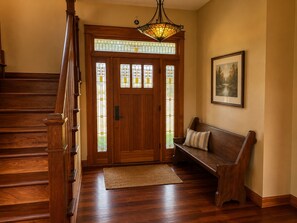 Una escalera de madera que conduce a una puerta con paneles de vidrio emplomado, un banco con cojín y un cuadro en la pared.