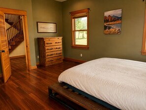 Un dormitorio con cama de madera, tocador, cuadro enmarco y ventana con vista a árboles.