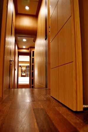 A corridor with wooden walls and a wooden floor, leading to a room with a bed and a window.