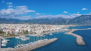 A coastal town with a marina, rocky breakwater, and mountains in the background.