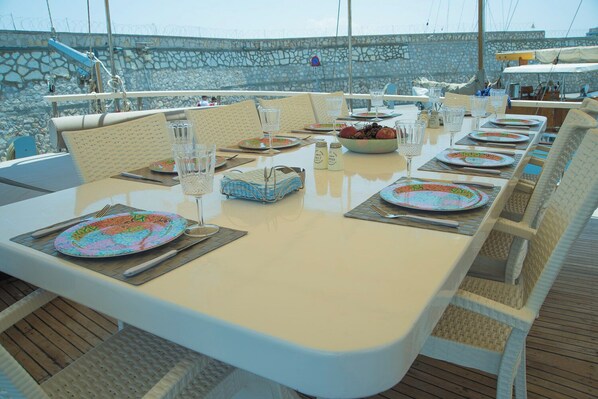 A dining table set with plates, cutlery, glasses, and a bowl of fruit on a deck with stone walls and a view of the sea.