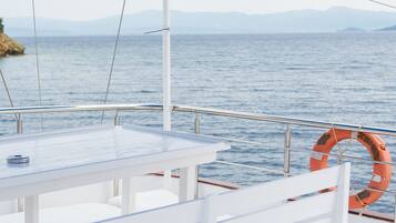 Un pont de bateau avec une table et des chaises, une bouée de sauvetage et une vue sur la mer et les montagnes lointaines.