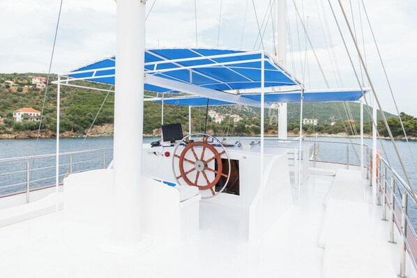 A boat deck with a steering wheel, a blue awning, and a view of a coastal area with houses.