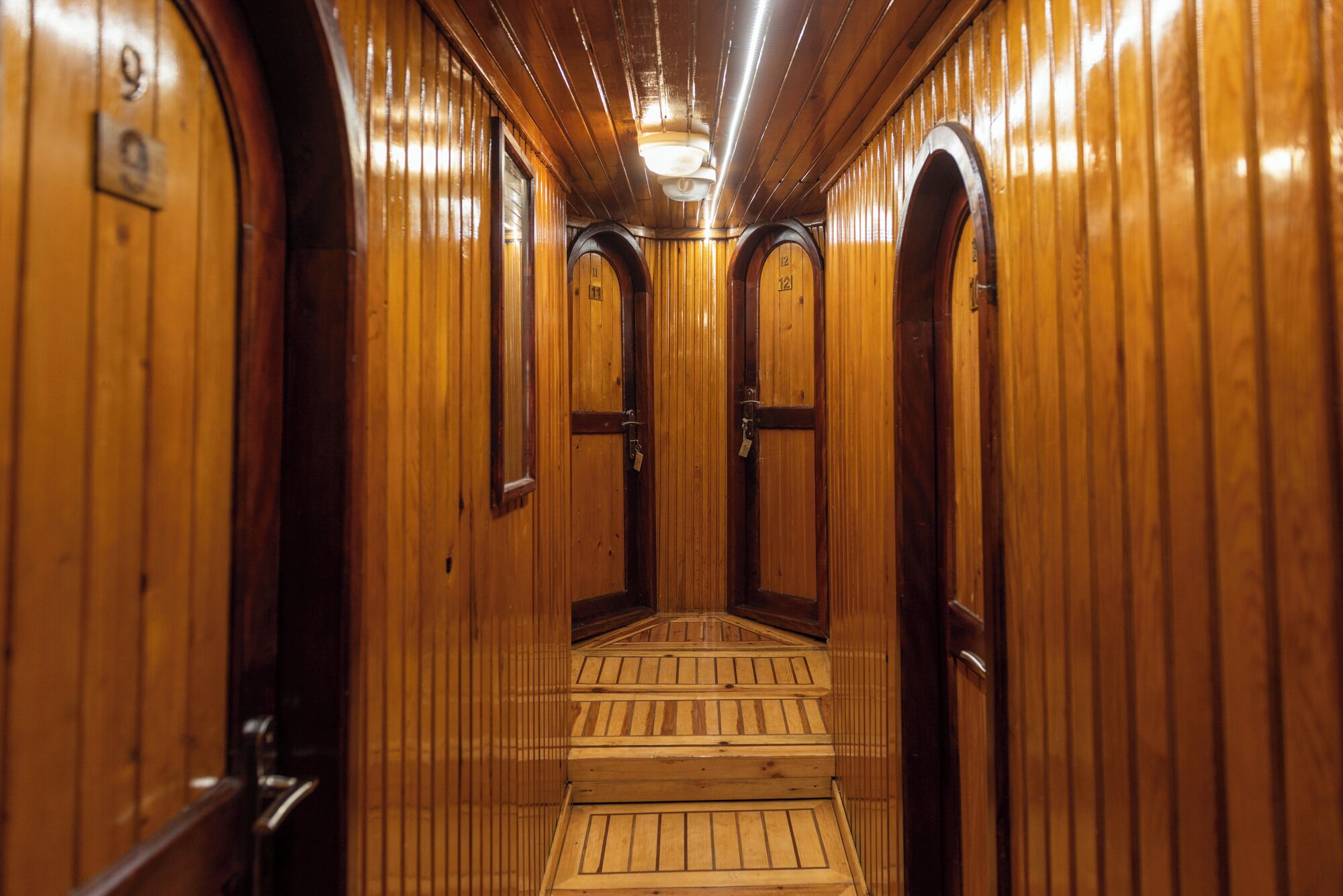 A wooden interior hallway with doors marked with numbers, a central staircase, and a ceiling light fixture.