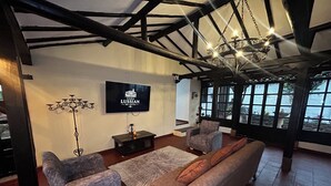 Un salon comprenant un canapé, deux fauteuils, un téléviseur, un lustre et une vue dégagée sur l’extérieur grâce à de grandes fenêtres.