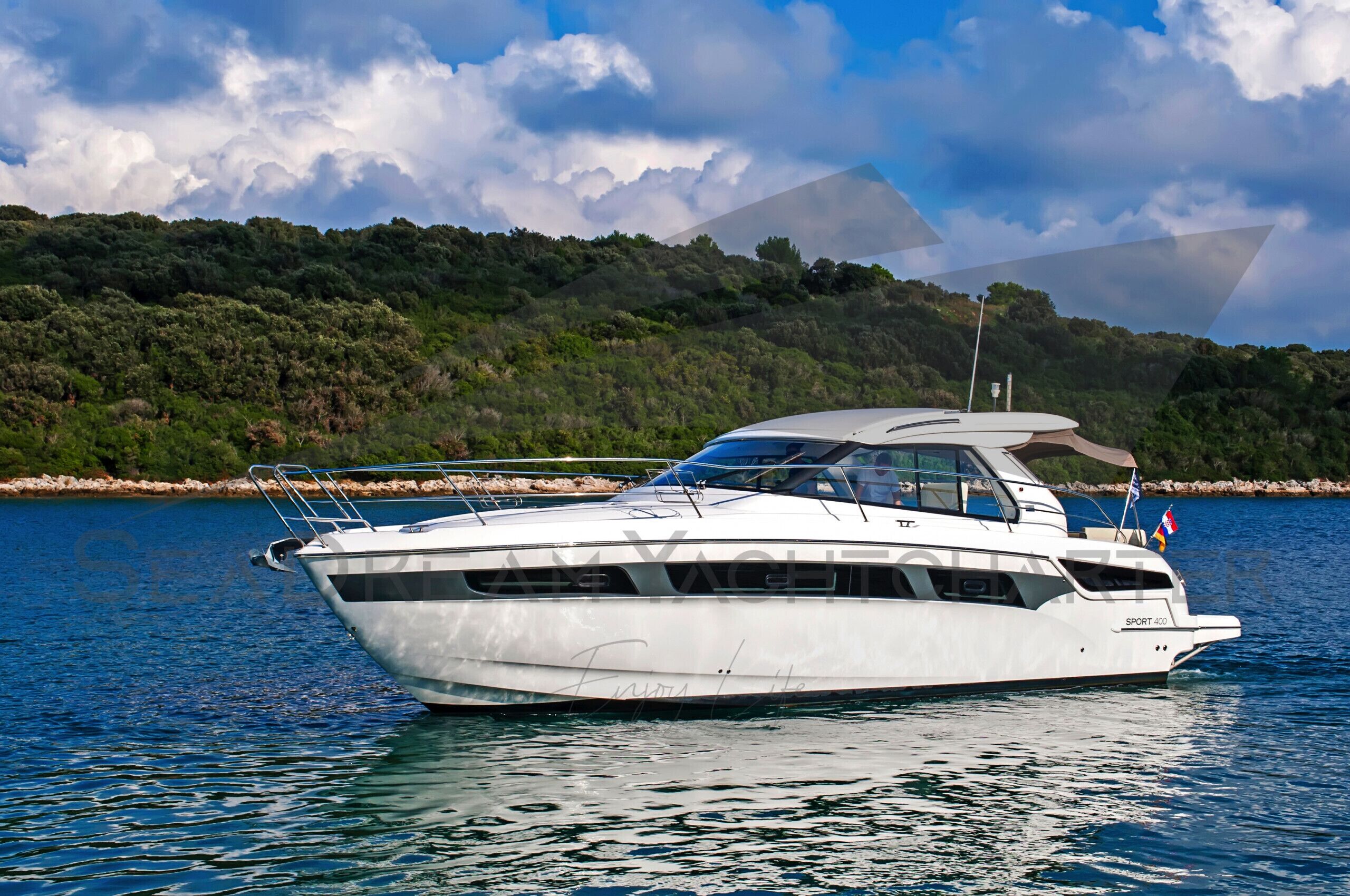Un yacht blanc, doté d’une cabine, d’un pavillon et d’un nom peint sur sa coque, est amarré dans une étendue d’eau, avec une colline boisée en arrière-plan.