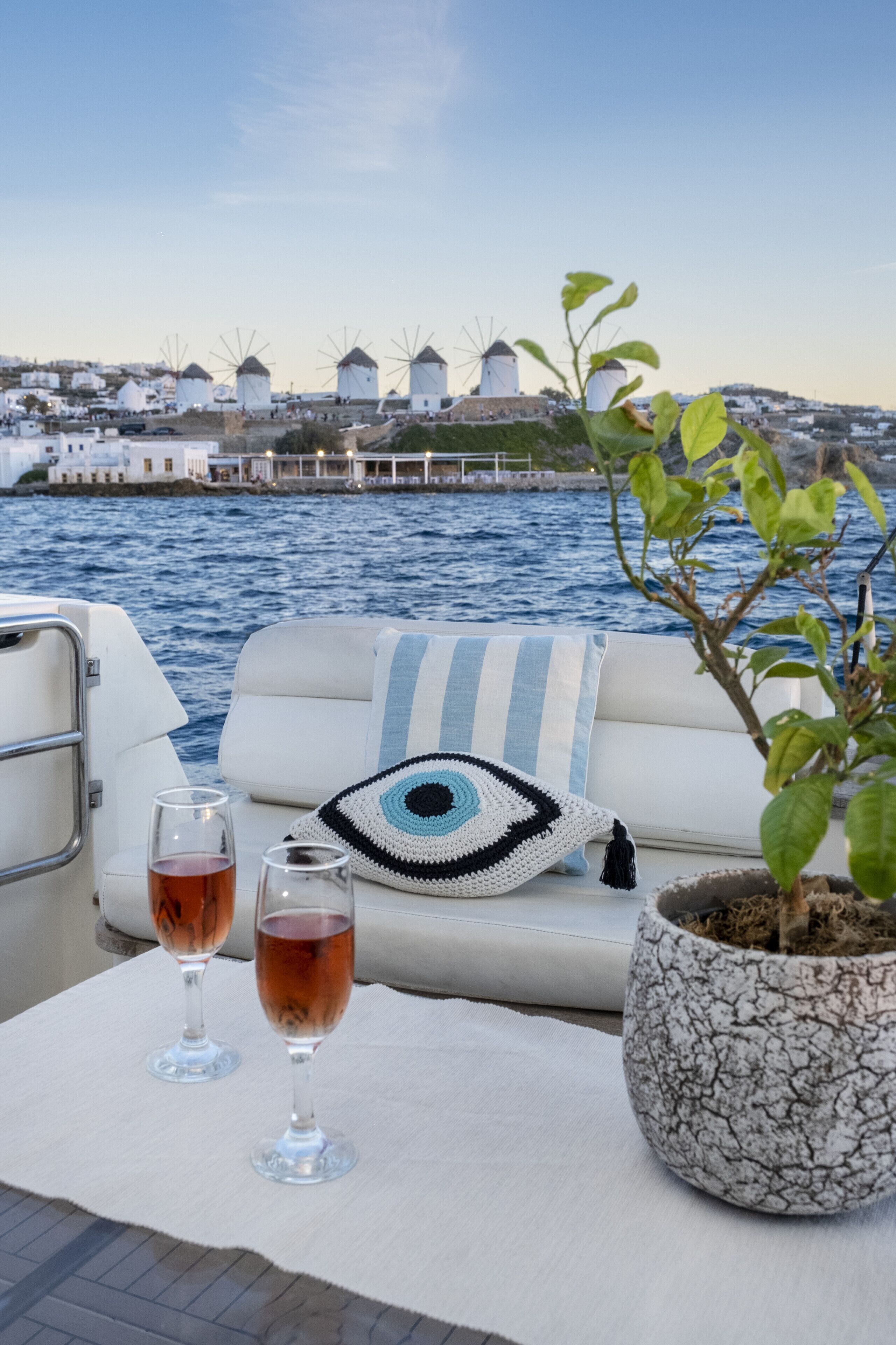 Cubierta de barco con una zona de estar blanca acolchada, dos copas de vino tinto, un cojín con motivos oculares de ganchillo y una planta en maceta.