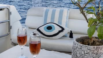 Cubierta de barco con una zona de estar blanca acolchada, dos copas de vino tinto, un cojín con motivos oculares de ganchillo y una planta en maceta.