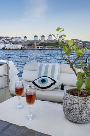 A boat deck with a white cushioned seating area, two glasses of red wine, a crocheted eye-patterned pillow, and a potted plant.