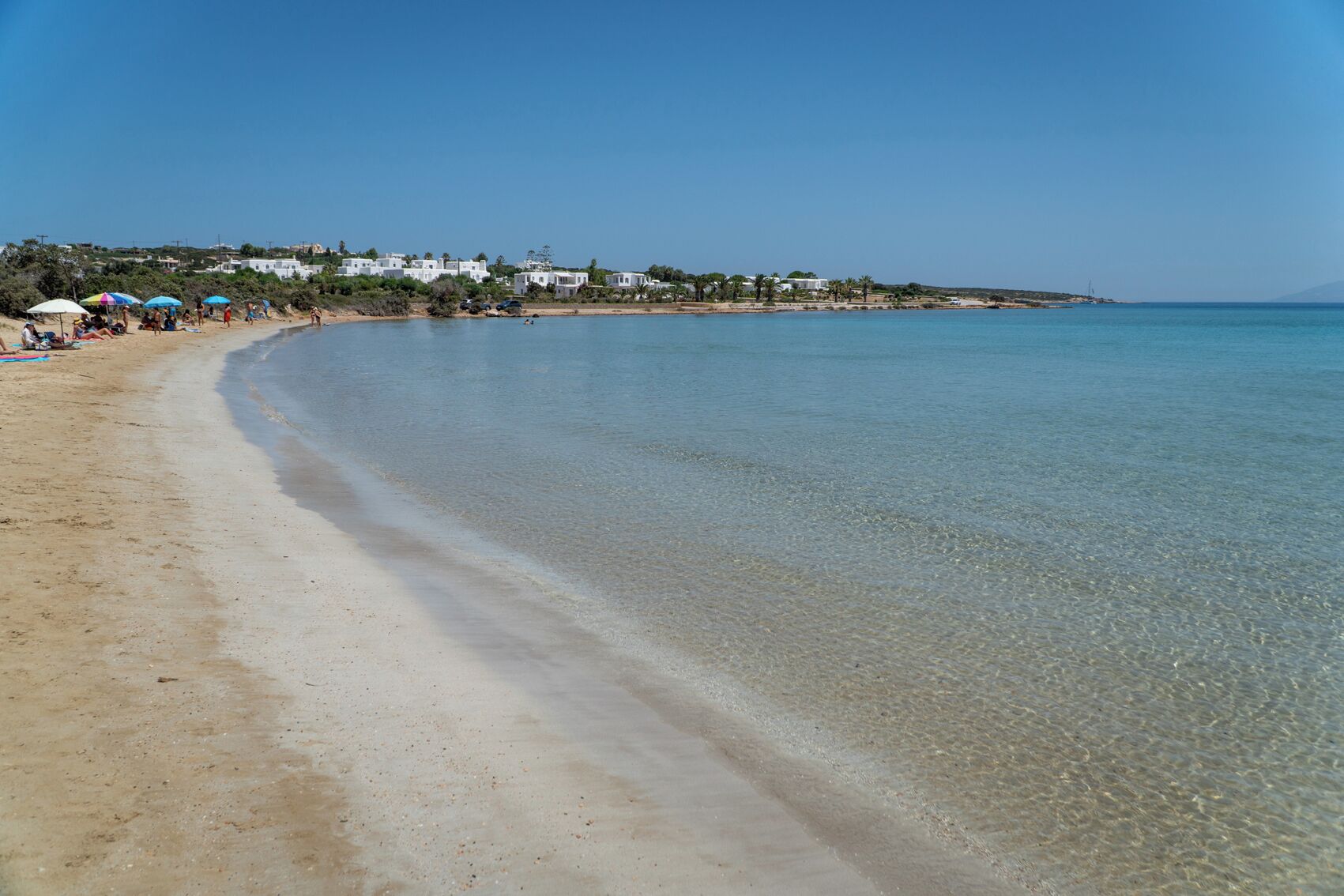 Una playa con agua cristalina, una orilla arenosa y edificios al fondo.