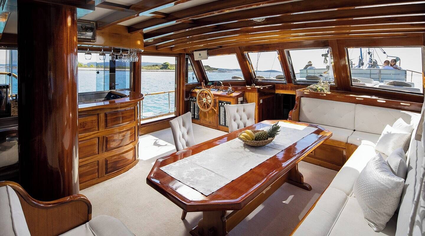 A boat interior with a dining table, cushioned seating, a wooden cabinet, and a steering wheel.