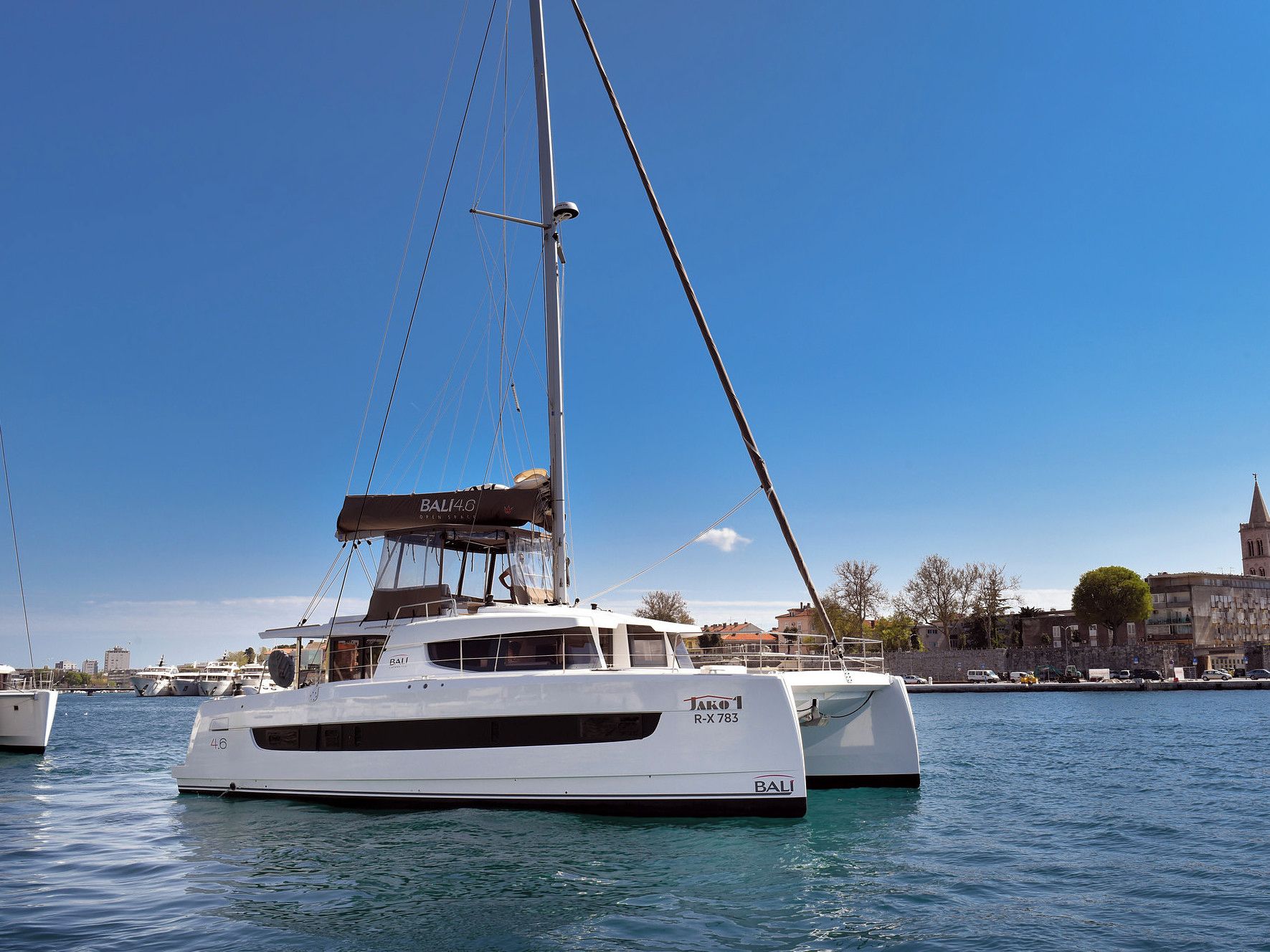 A white yacht named 'BALI 4.6' docked at a marina with a clear blue sky and a cityscape in the background.