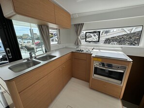 Une cuisine compacte équipée d’un évier, d’une cuisinière et de placards, avec de grandes fenêtres offrant une vue sur un port de plaisance.