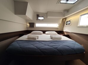 Un lit avec une couette bleue, des oreillers blancs et des serviettes enroulées. Il y a une petite fenêtre avec des rideaux et un téléviseur accroché au mur.