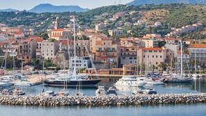 Un port de plaisance où sont amarrés des bateaux, entouré d’une ville avec ses bâtiments et dominé par un paysage montagneux.