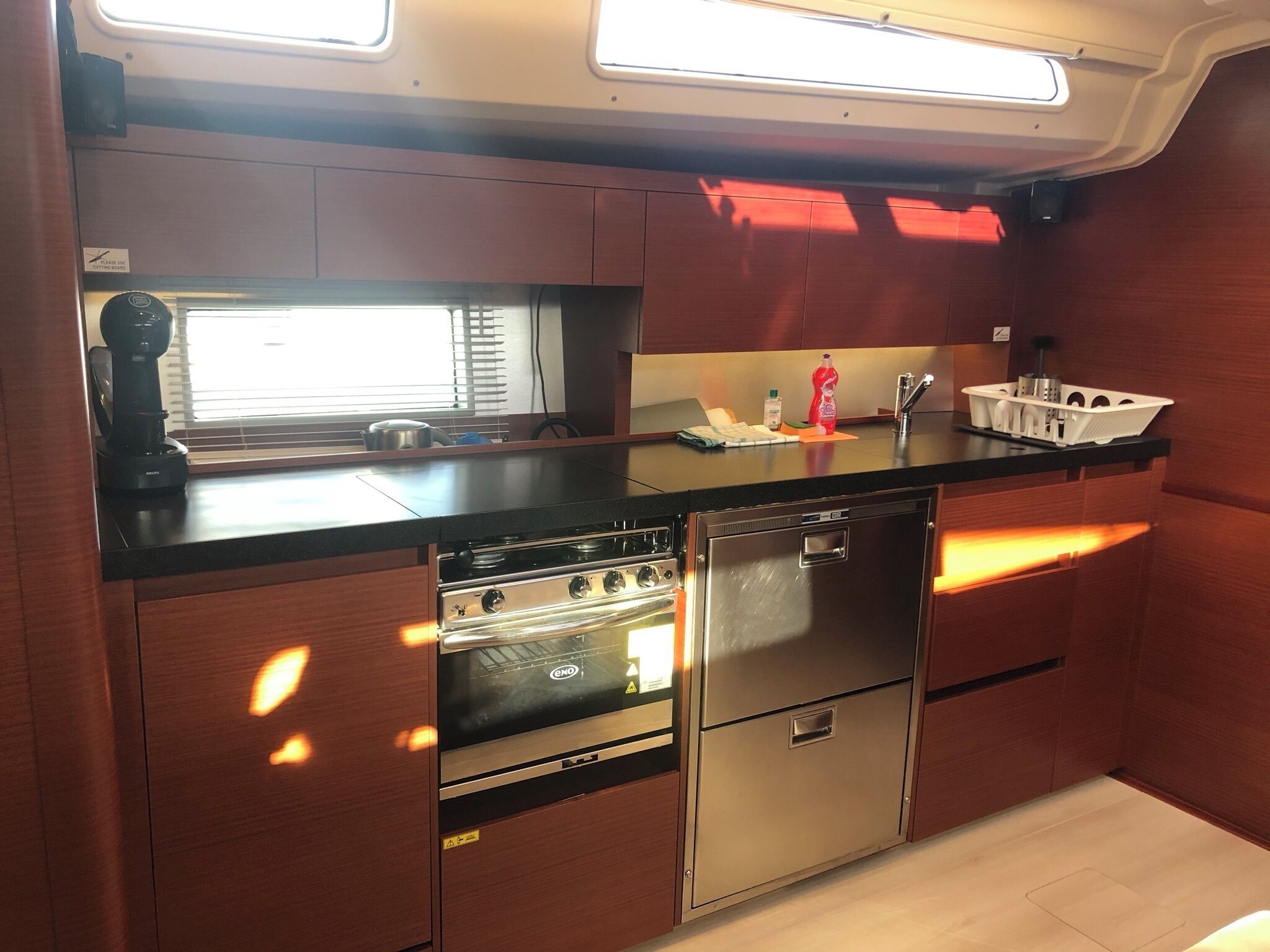 A compact kitchen with a stove, oven, and dishwasher, wooden cabinets, and a window with blinds.
