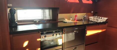 A compact kitchen with a stove, oven, and dishwasher, wooden cabinets, and a window with blinds.