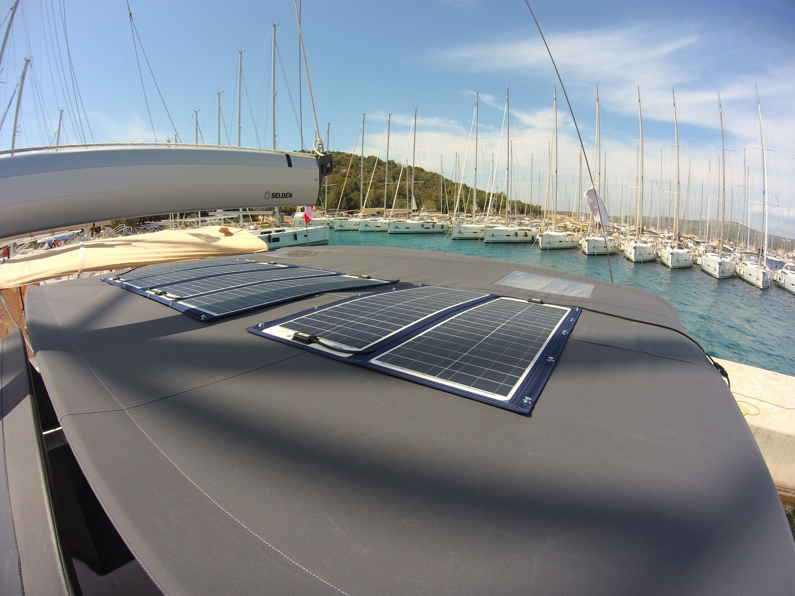 Cubierta de barco con paneles solares, un puerto deportivo con veleros y un paisaje montañoso al fondo.