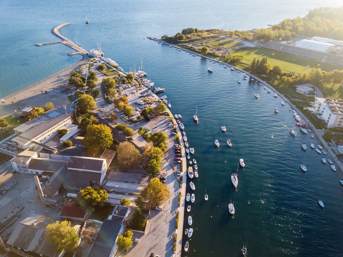 An aerial view of a coastal marina with boats docked along the shore, a curved breakwater, and a mix of residential and commercial buildings.