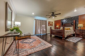 A bedroom with a wooden bed, a nightstand, a desk with a lamp, a large mirror, and a ceiling fan. - Casa de Valdes: Spacious Two-Story Home Near SeaWorld and Military Bases (San Antonio)