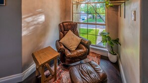A leather armchair with a cushion, a small wooden table, and a potted plant by a window with a view of a lawn and a house. - Casa de Valdes: Spacious Two-Story Home Near SeaWorld and Military Bases (San Antonio)