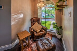 A leather armchair with a cushion, a small wooden table, and a potted plant by a window with a view of a lawn and a house. - Casa de Valdes: Spacious Two-Story Home Near SeaWorld and Military Bases (San Antonio)
