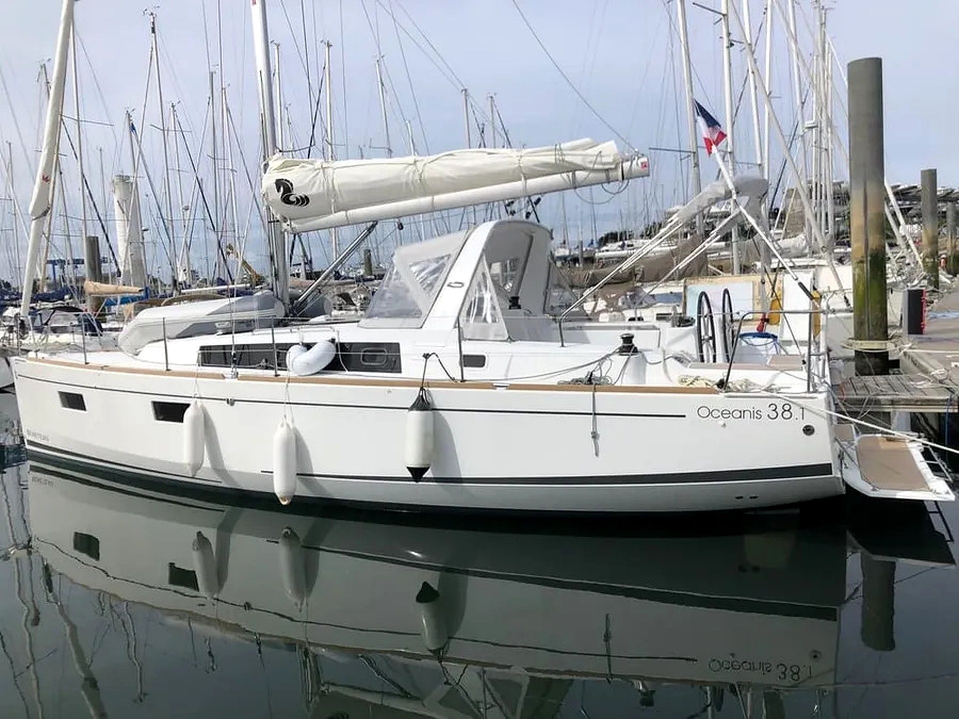 Un yacht blanc, baptisé « Oceanis 38.1 », était amarré dans une marina dotée de plusieurs mâts et arborant un pavillon.