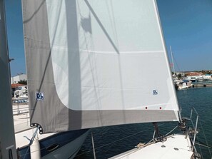 Un voilier à voile blanche, avec un logo sur la voile, et un port de plaisance avec des bateaux en arrière-plan.