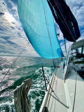 A sailboat with a large sail and ropes on deck, set against a clear sky and ocean view.