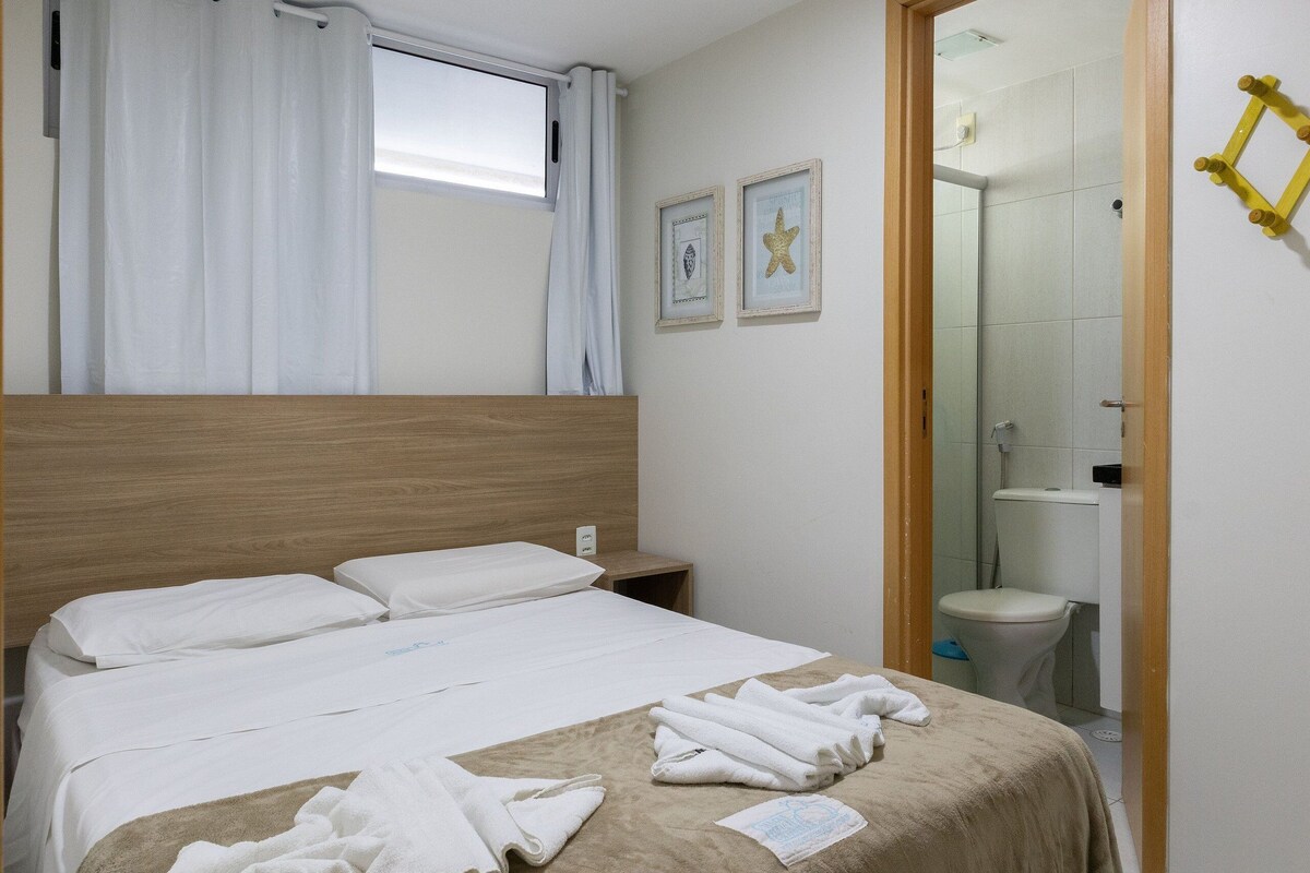 A hotel room with a bed, a nightstand, a window with curtains, a bathroom with a toilet and shower, and framed pictures on the wall.