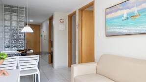 Ein modernes Wohnzimmer mit einem beigefarbenen Sofa, einem Essbereich mit weißen Stühlen, einer Küche mit Glasabtrennung und einem Gemälde von Booten an der Wand.