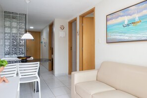 A modern living room with a beige sofa, a dining area with white chairs, a kitchen with a glass partition, and a painting of boats on the wall. - Ocean Residence (Ipojuca)