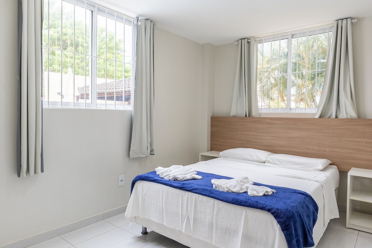 A hotel room with a bed, a nightstand, a window with curtains, and a view of palm trees outside.