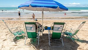 Ein Strand mit zwei grünen Liegestühlen unter einem blau-weiß gestreiften Sonnenschirm, im Hintergrund das Meer.