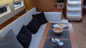 Interior de un barco con un banco acolchado blanco, una mesa con un cuenco y vasos, y una pared de madera con ventanas.