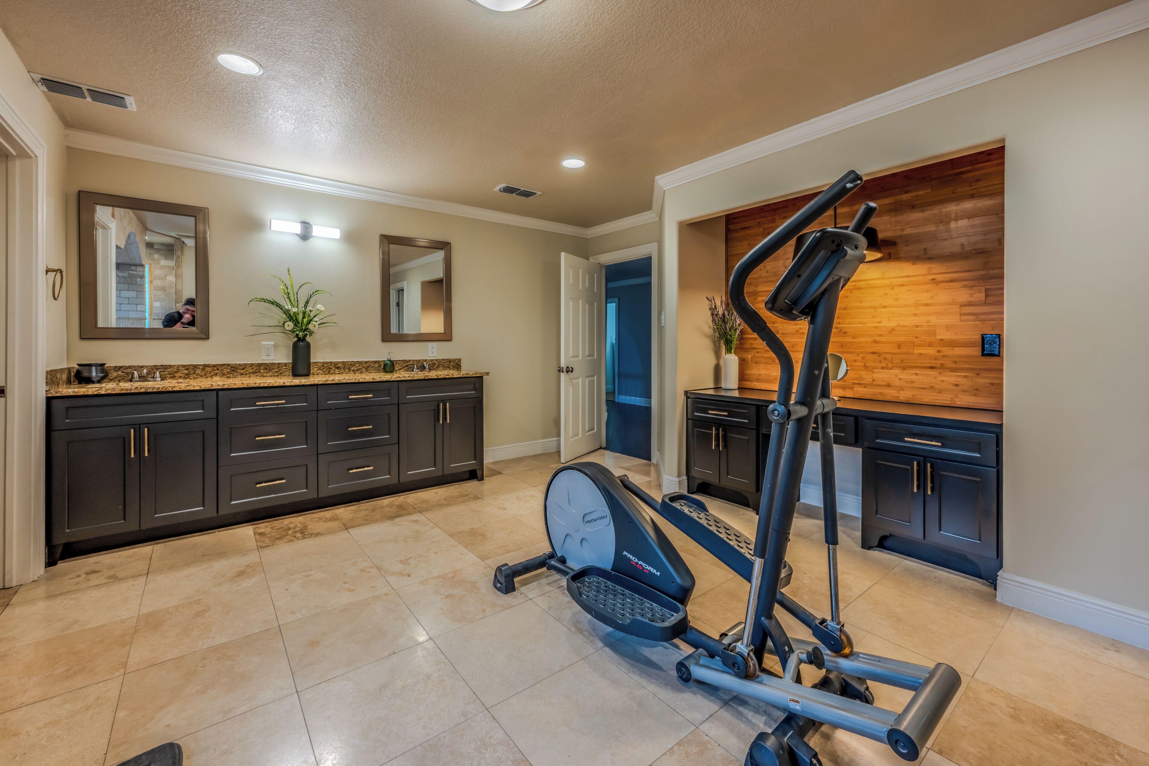 Une salle de sport à domicile équipée de matériel d’exercice, un coin cuisine avec des meubles sombres et un mur d’accent en bois, et une salle de bain avec un grand miroir et des plantes en pot.