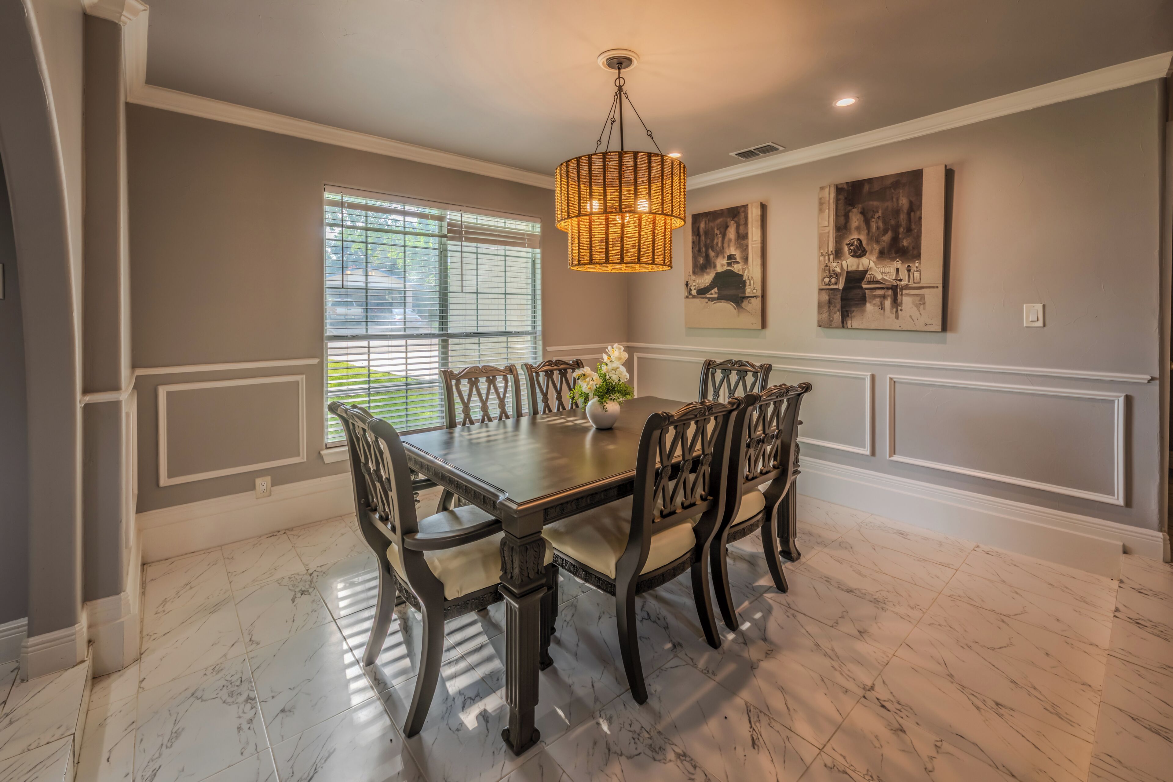 A dining room with a marble floor, a wooden table, and chairs, a hanging light fixture, and artwork on the walls.