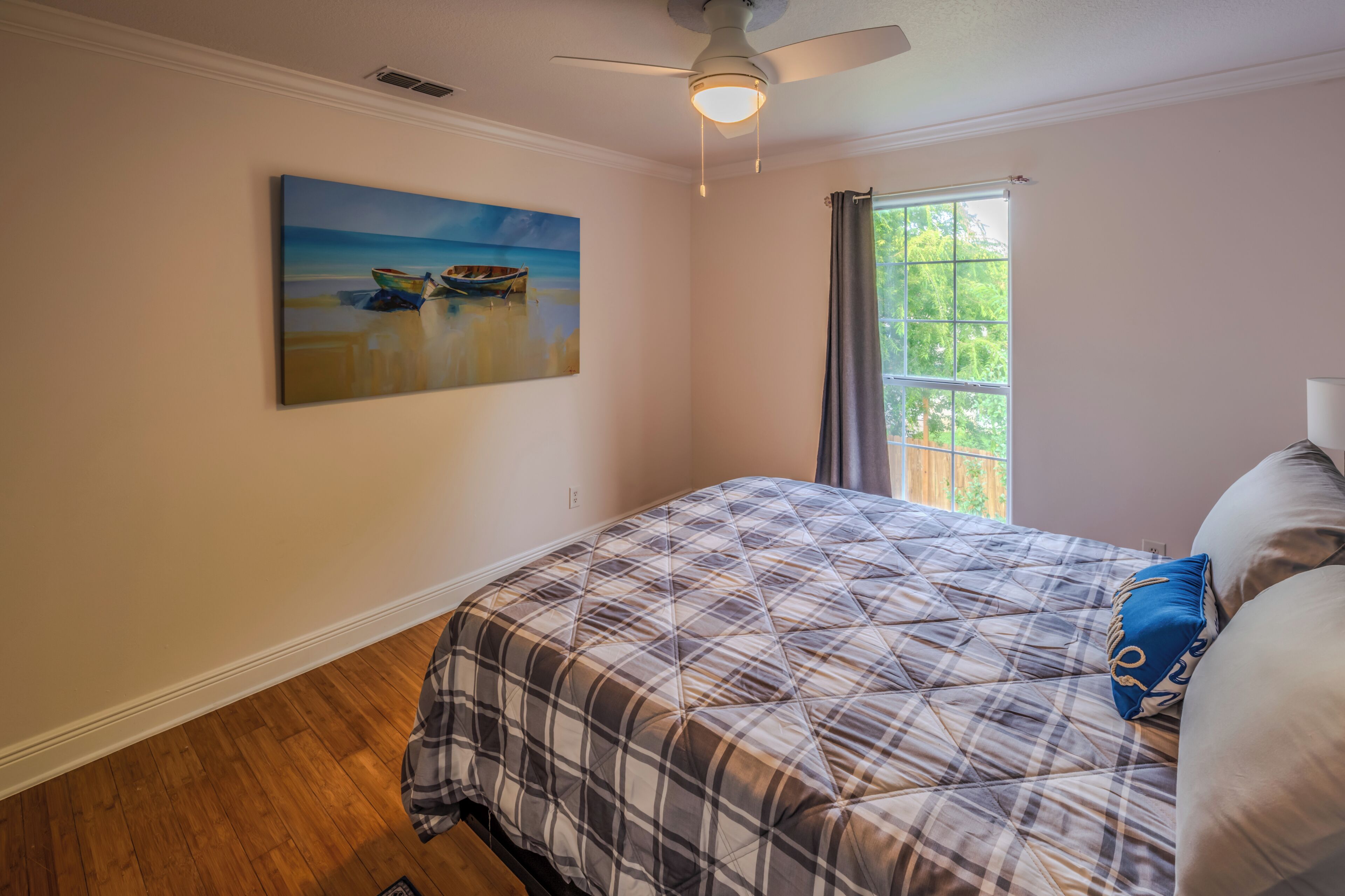 Een slaapkamer met een bed, een plafondventilator, een schilderij van boten aan de muur en een raam met gordijnen.