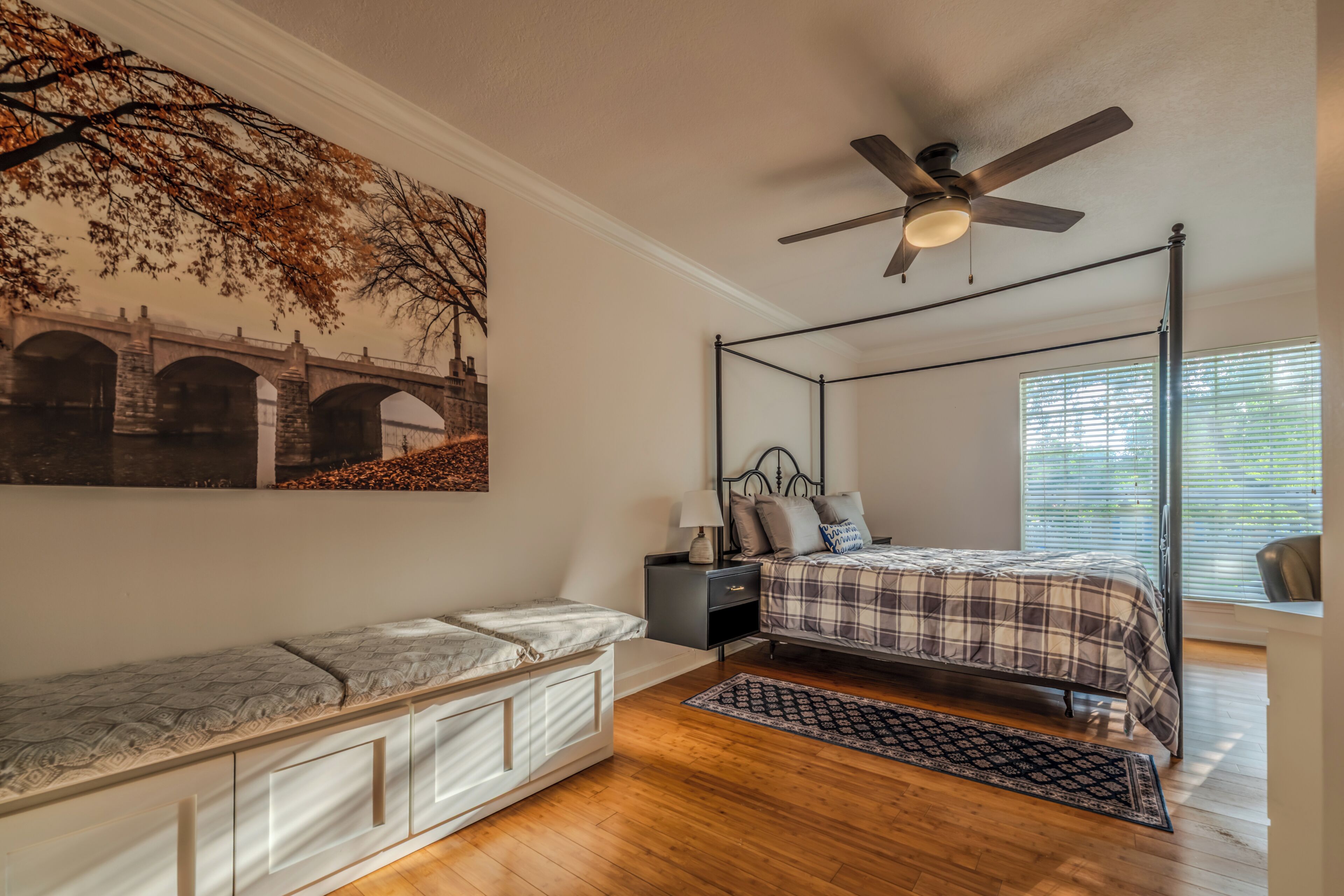A bedroom with a four-poster bed, a nightstand, a ceiling fan, and a large window.