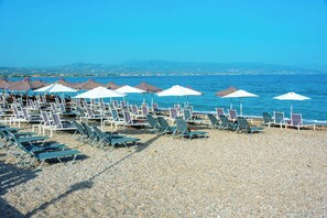 A beach with rows of sun loungers and umbrellas, a pebble beach, and a clear blue sea.