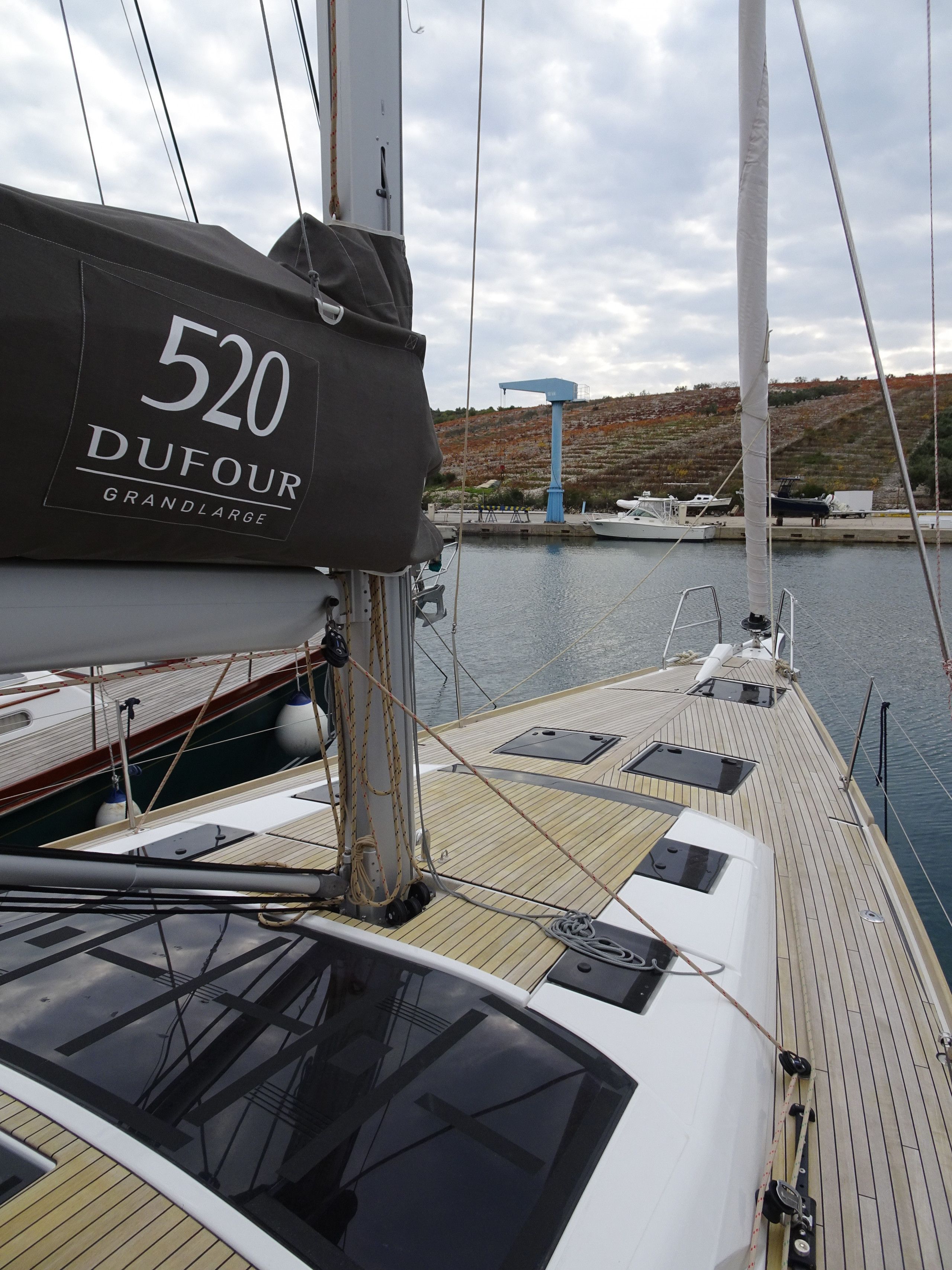 Eine Segelyacht liegt in einer Marina festgemacht, mit einer '520 DUFOUR GRANDLARGE'-Abdeckung, einem Holzboden und Blick auf Wasser und Hügel im Hintergrund.