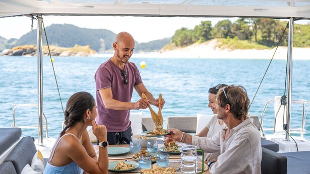 Een eettafel met borden eten, glazen en een karaf op een boot met uitzicht op zee en een strand.