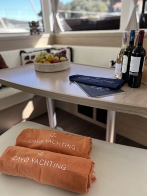 A table with a bowl of fruit, bottles of wine, a book, and towels with text. - Sun-Kissed Sailing Adventure in the Ionian Islands, Lefkada (Lefkada)