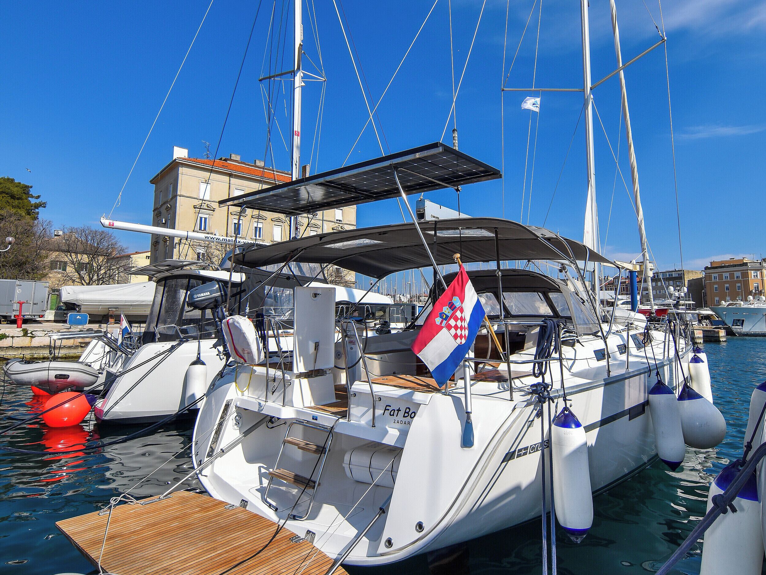 Un voilier est amarré dans une marina, on peut voir un drapeau, un pont couvert et un bâtiment en arrière-plan.