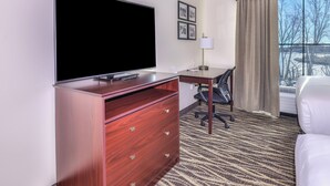 A hotel room with a flat-screen TV, a desk with a chair, a large window with a view of trees, and a bed. - Cobblestone Hotel & Suites - Montevallo (Montevallo)
