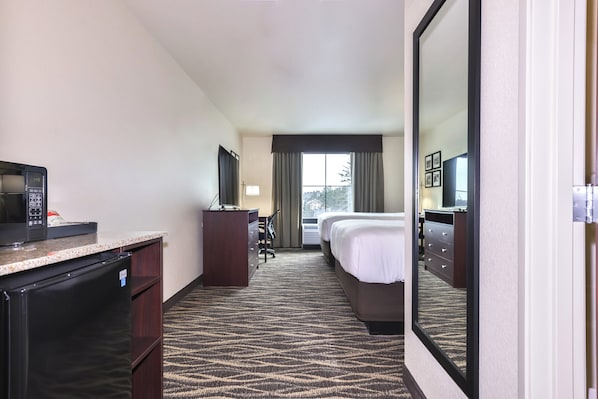 A hotel room with a kitchenette, a bed, a desk, a chair, a mirror, and a window with curtains.