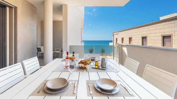 Una mesa de comedor dispuesta con platos, cubiertos y vasos, con vista al mar y edificios al fondo.