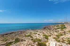 A rocky coastline with a clear blue sky, a paved walkway, and a distant view of buildings. - Zefiro Suite by BarbarHouse (Gallipoli)